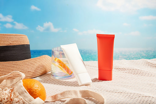 Sunscreen tubes in different colors displayed on the scarf with a water cup, beach hat and a mesh bag of oranges. Cosmetic concept with sun protection and regenerating products for skin