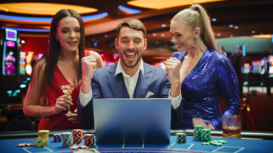 Portrait of a Happy Man with Two Female Partners Betting Online on a Laptop Computer while Sitting in a Luxury Casino. Lucky Man and Glamorous Women Celebrating Blackjack Winning Jackpot .