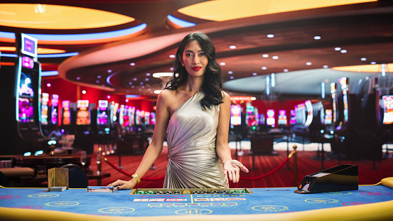 Portrait of a Professional Female Croupier Looking at the Camera and Welcoming You to a Table. Beautiful Asian Dealer in Silver Dress Dealing Playing Cards in a Casino for a Game of Baccarat