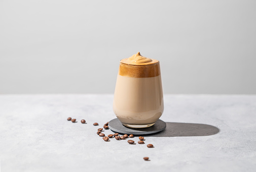 Dalgona coffee. Whipped instant coffee in a glass with beans and shadow on a light background.  The concept of a trendy and popular drink. Free space for text.