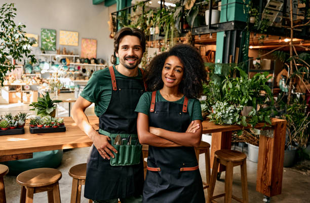 Small flower business. Front view of caucasian man and african american woman in aprons smiling at camera white standing at own shop. Happy couple selling fresh green plants for home decor. small business stock pictures, royalty-free photos & images