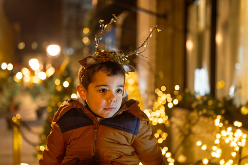 Cute little boy with Christmas ornaments. He is outdoor, the city is Christmas decorated