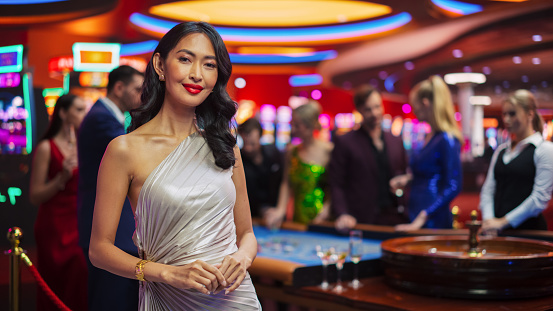 Advertising Campaign Template for an Online Casino and Betting Website with an Asian Woman Posing, Looking at Camera, Pointing to CTA Placeholder in a Glamorous Casino with Gamblers Playing Roulette