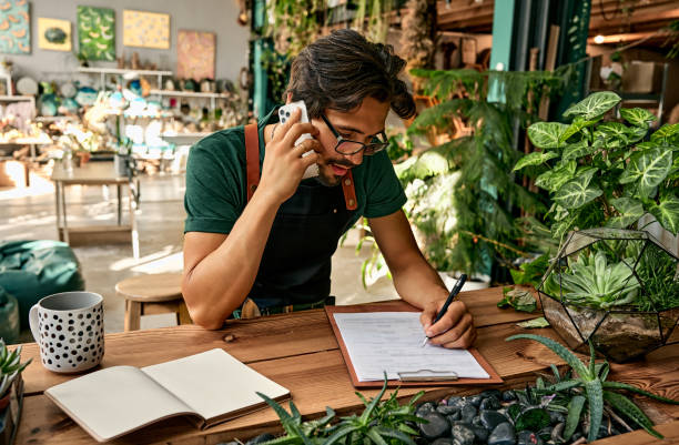 Service and shopping concept. Handsome caucasian man in eyeglasses and apron making notes on clipboard during mobile conversation at flower shop.