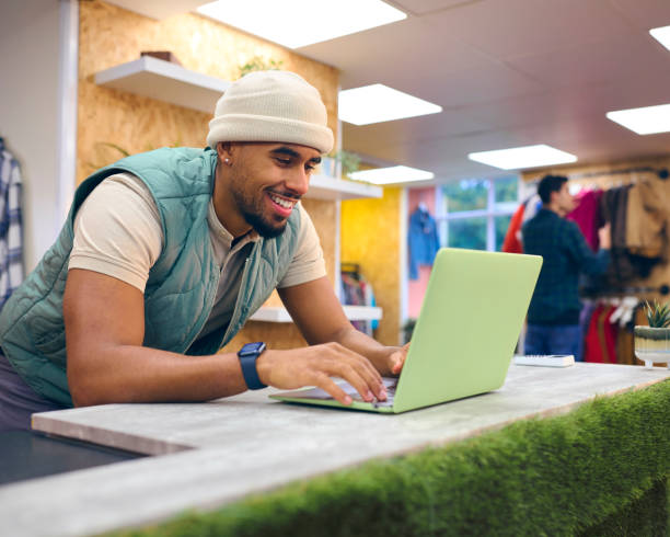 Assistente alle vendite maschile alla cassa in un negozio di moda o abbigliamento pop-up utilizzando il laptop - foto stock