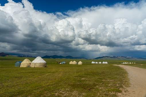 A serene nomadic settlement of circular yurts unfolds along the verdant plains of Son-Kul, embraced by the grandeur of distant mountain silhouettes and a canvas of sprawling, cloud-laced blue skies.
