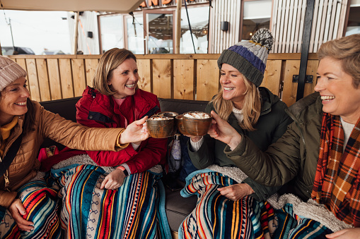 Four women are sitting wrapped in colourful blankets at an outside cafe drinking hot chocolate with cream on. They are toasting each other and smiling.
