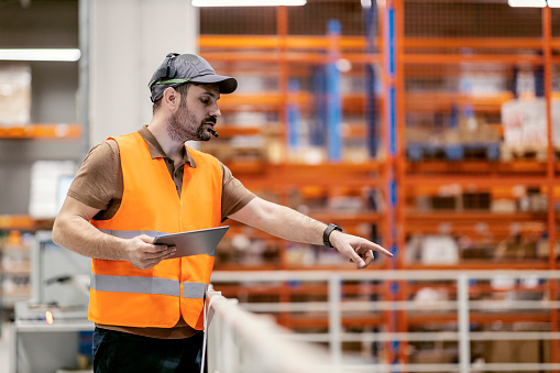 A logistics worker is controlling and giving orders to workers from distance with use of headset and tablet.