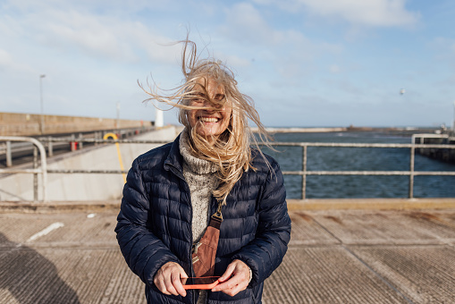 A woman stands on a harbour looking at the camera, her hair is blowing across her face. She is holding a red mobile phone.