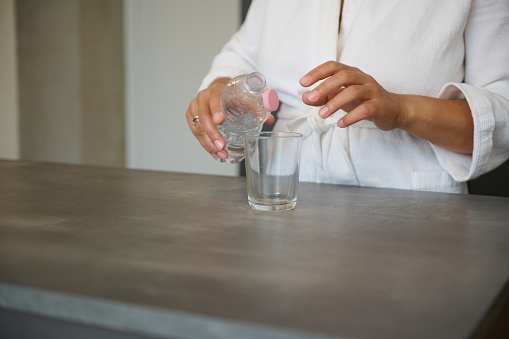 Close-up hands of an unrecognizable woman in white waffle robe holding glass of refreshing water, standing at kitchen island in modern minimalist home interior. Body care. Healthy habits and lifestyle