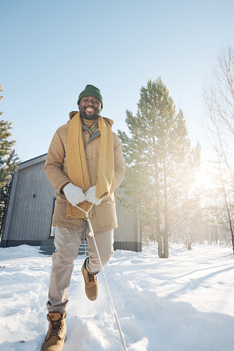 Young cheerful African American man in winterwear pulling rope of sledge and running down snow path on sunny and frosty winter day