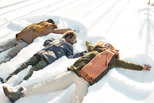 Three cheerful intercultural people in winterwear lying in snowdrift while playing and having fun at leisure or during winter vacation