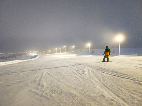 Rear view of a snowboarder enjoying the slope under artificial lights during the polar night in Lapland, Finland, Scandinavia