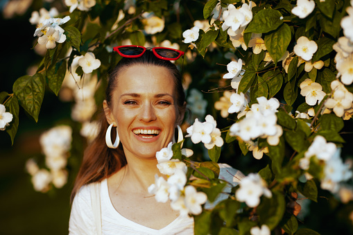 Summer time. Portrait of happy stylish woman in white shirt near flowering tree.