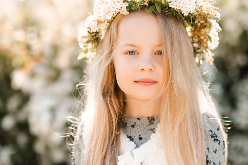 Cute kid blonde girl 3-4year old wear floral wreath posing over flowers at background close up. Looking at camera. Spring season. Childhood.