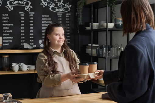 Female clerk with Down syndrome in apron passing two disposable cups of coffee to guest of cafe over counter while serving clients