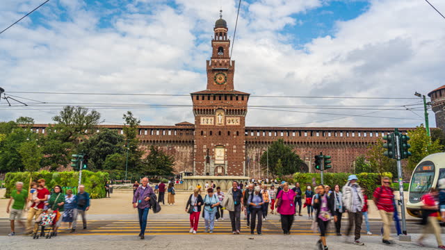 Time lapse of Crowd of People tourist pedestrian walking and crosswalk with moving tram on city street traffic road intersection at Filarete Tower and Sforzesco Castle in summer, central Milan, Italy