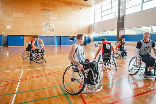 Wide angle view of male wheelchair basketball players in 20s and 30s moving around court during practice game.