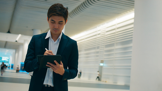 Smart young Asian businessman CEO wearing business suit using digital tablet, working, writing on note pad and standing in office, Professional executive manager working on tech device, Business people with smart technology and social network