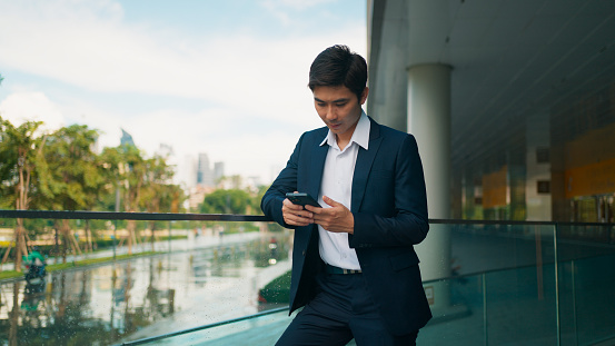 Smart young Asian businessman using smartphone, working, reading news and chatting with people by online message at beautiful modern office building, Business people with smart technology social network, Surfing internet over mobile phone device