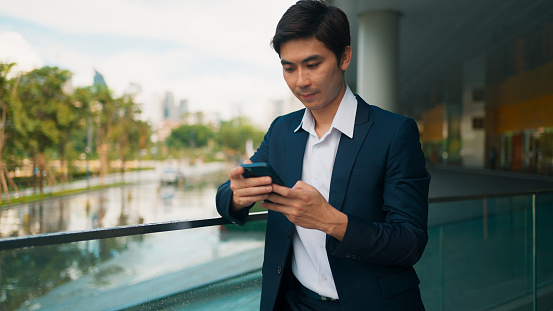 Smart young Asian businessman using smartphone, working, reading news and chatting with people by online message at beautiful modern office building, Business people with smart technology social network, Surfing internet over mobile phone device