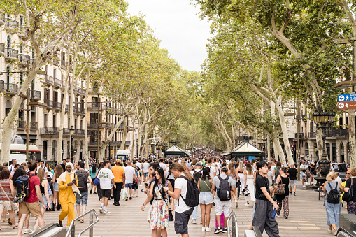 Barcelona, Spain - Aug 4, 2023: Tourists walk past sidewalk cafes on the famous La Rambla street through the historic Mediterranean city of Barcelona, Spain.