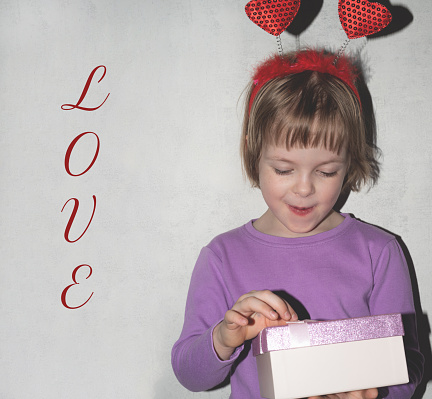 Happy surprised child girl in a headband with hearts looks at a box with a gift. Text Love in the background. Valentine's day holiday concept