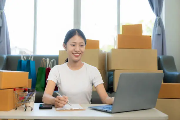 Photo of Small business startup, SME owner, young Asian woman checking online orders, selling products, working with boxes, freelancer working at home using laptop, packaging, delivery concept.