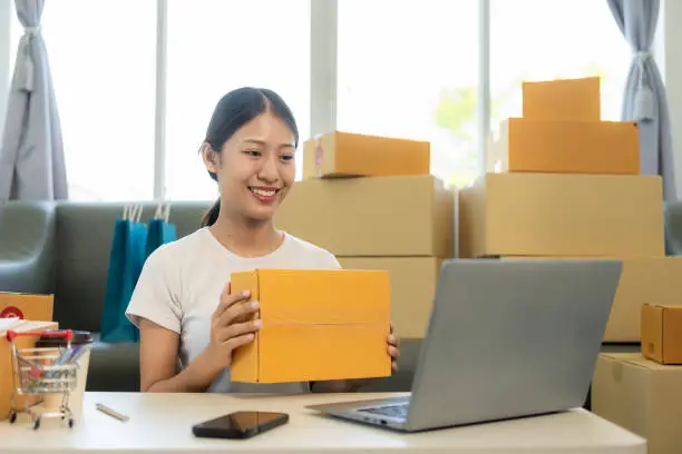 Photo of Small business startup, SME owner, young Asian woman checking online orders, selling products, working with boxes, freelancer working at home using laptop, packaging, delivery concept.