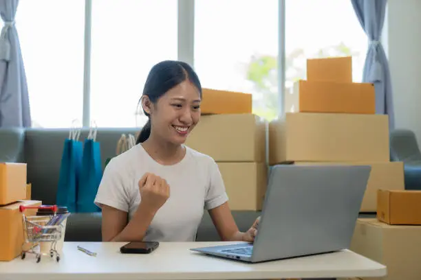 Photo of Small business startup, SME owner, young Asian woman checking online orders, selling products, working with boxes, freelancer working at home using laptop, packaging, delivery concept.