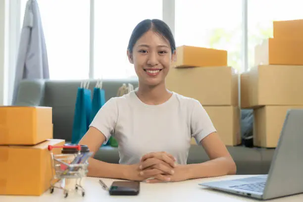 Photo of Small business startup, SME owner, young Asian woman checking online orders, selling products, working with boxes, freelancer working at home using laptop, packaging, delivery concept.