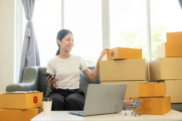 Photo of Small business startup, SME owner, young Asian woman checking online orders, selling products, working with boxes, freelancer working at home using laptop, packaging, delivery concept.