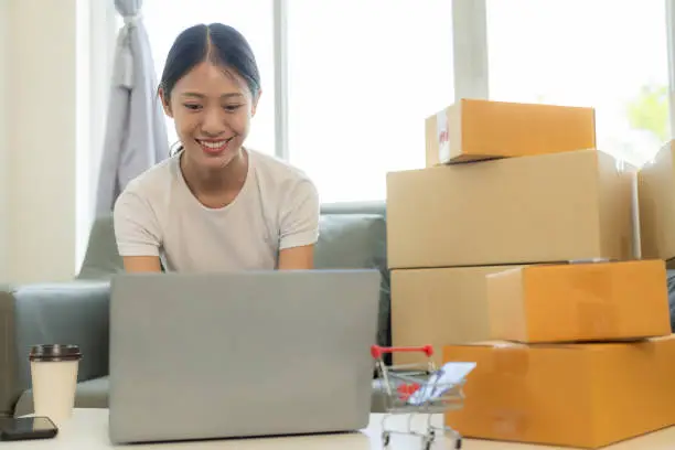 Photo of Small business startup, SME owner, young Asian woman checking online orders, selling products, working with boxes, freelancer working at home using laptop, packaging, delivery concept.