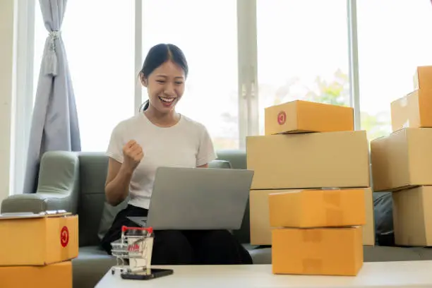 Photo of Small business startup, SME owner, young Asian woman checking online orders, selling products, working with boxes, freelancer working at home using laptop, packaging, delivery concept.