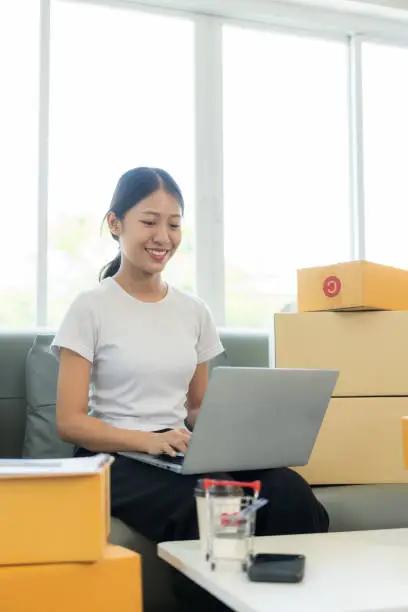 Photo of Small business startup, SME owner, young Asian woman checking online orders, selling products, working with boxes, freelancer working at home using laptop, packaging, delivery concept.