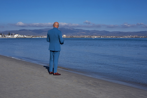 Elegant gentleman in a blue suit, strolling on Poetto Beach: a stylish contrast of formal attire against the serene coastal backdrop