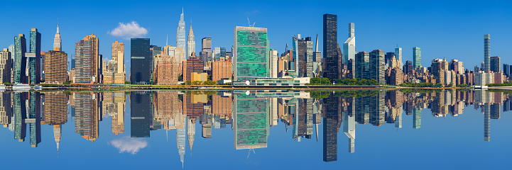 High Resolution Stitched Panorama of New York Skyline with UN Building (Headquarters of the United Nations), Chrysler Building, Empire State Building, Manhattan Upper East Side Residential and Office High-rises, FDR drive, Green Trees and Morning Clear Blue Sky all Reflected in Water of East River. Canon EOS 6D (Full Frame censor) DSLR and Canon EF 85mm f/1.8 lens. 3:1 Image Aspect Ratio. This image is downsized to 50MP. The Original image resolution is 124MP or 19,314 x 6,438 px.