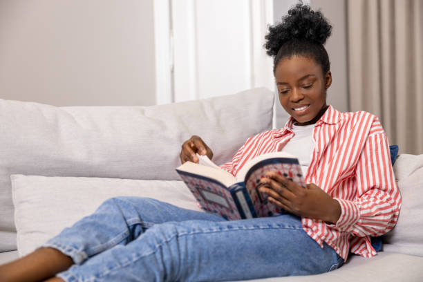 istock Black woman reading book while relaxing on couch in home interior. 1924942649