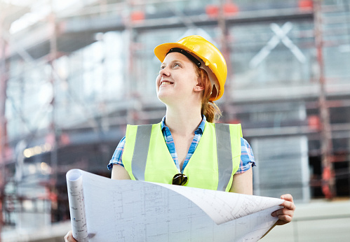 Contended, happy young construction worker on site, looking up as she holds a large paper blueprint.