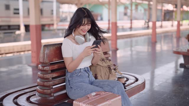 Asian female tourist waiting for a train at the railway station. She's using social media or chatting with friends on her smartphone. Ideal for travel.