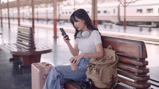 Asian female tourist waiting for a train at the railway station. She's using social media or chatting with friends on her smartphone, enjoying a cup of coffee