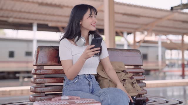 Asian female tourist waiting for a train at the railway station. She's using social media or chatting with friends on her smartphone, enjoying a cup of coffee