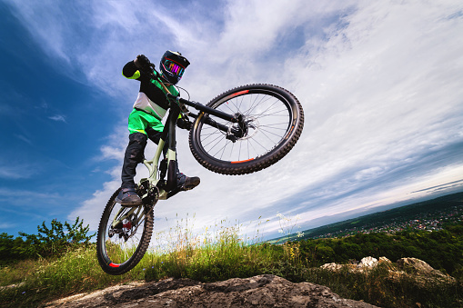 A professional racer jumps on a bicycle against a blue cloudy sky. Sunny summer day. Low angle view of a man on a mountain bike jumping in the mountains