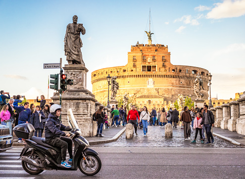 Rome, Italy - Traffic passing on the street as tourists cross Ponte Sant’Angelo and photograph Castel Sant'Angelo at sunset.
