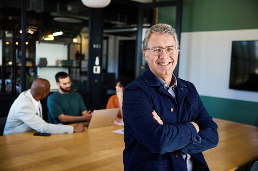 Portrait of a smiling mature businessman standing with his arms crossed in an office boardroom with colleagues talking in the background
