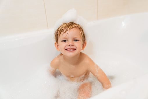 Cute little boy with foam hat taking a bath in the tub and looking at camera. Space for text