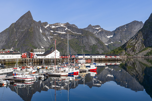 The tranquil harbor of Hamnoy, enveloped by towering cliffs, showcases a pristine reflection of boats and traditional Norwegian buildings. Lofoten Island, Norway