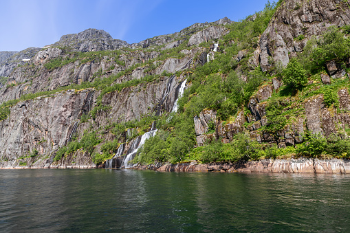 Multiple waterfalls cascade down the mossy cliffs of Trollfjorden in Lofoten, Norway, merging with the fjord's waters beneath a sunny sky.