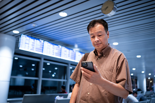 Mature Asian male business man checking his electronic boarding pass using smart phone while waiting for the flight at the airport departure hall.