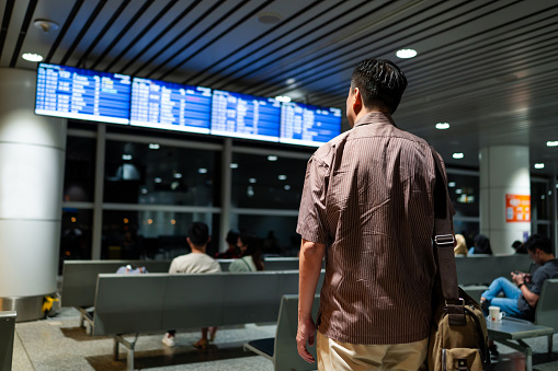 Asian male business traveler in smart casual outfit checking boarding time and waiting in front the arrival departure board.